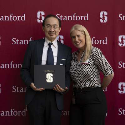 Two individuals pose for a photo holding a certificate in front of a Stanford-themed backdrop during an event.