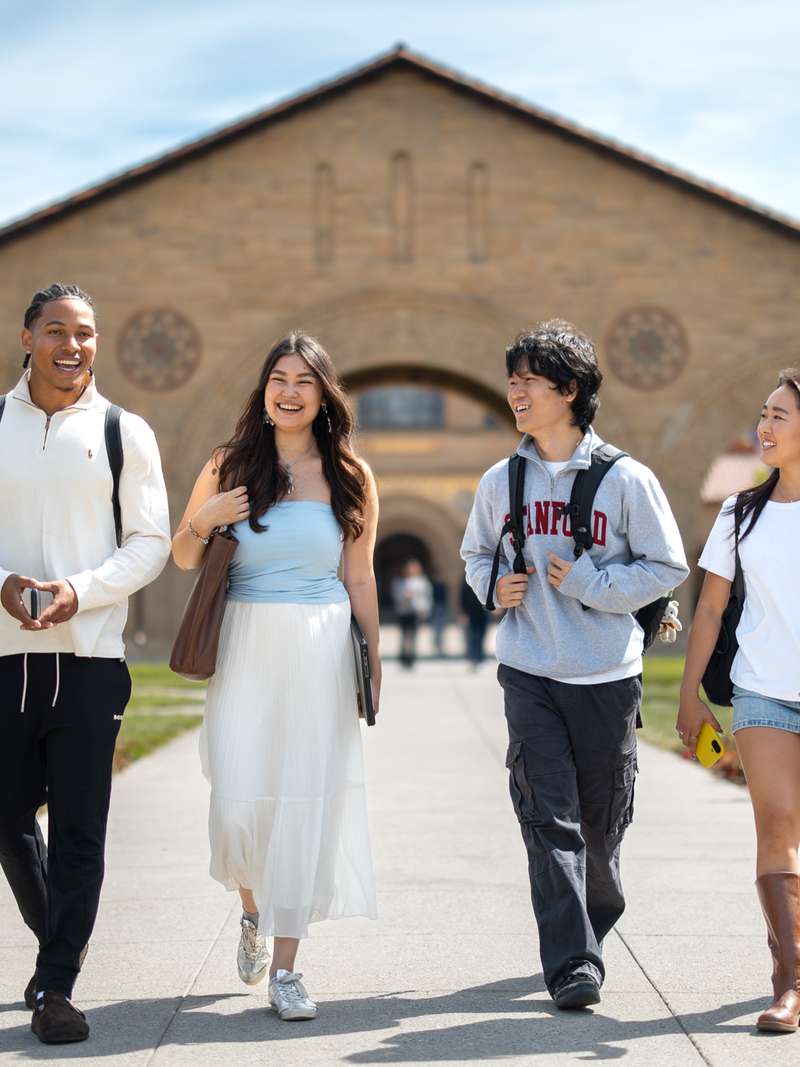Four students walk together on a campus path lined with flowers, with a large, historic building in the background.