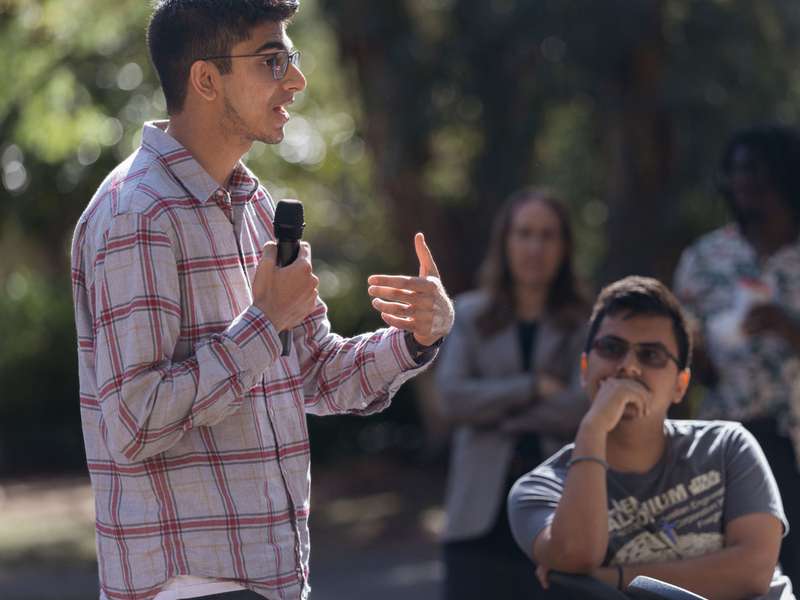A student speaks into a microphone at an outdoor campus event while others listen attentively in the background.