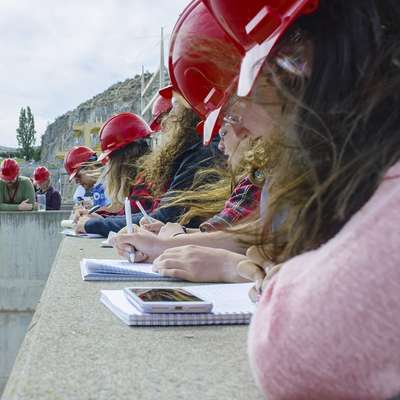 Students in red hard hats take notes while observing a river and surrounding landscape during a field study.