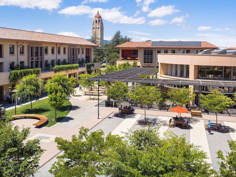 A sunny courtyard on the Stanford campus near the CEMEX Auditorium with seating areas and trees, and the Hoover Tower visible in the background.