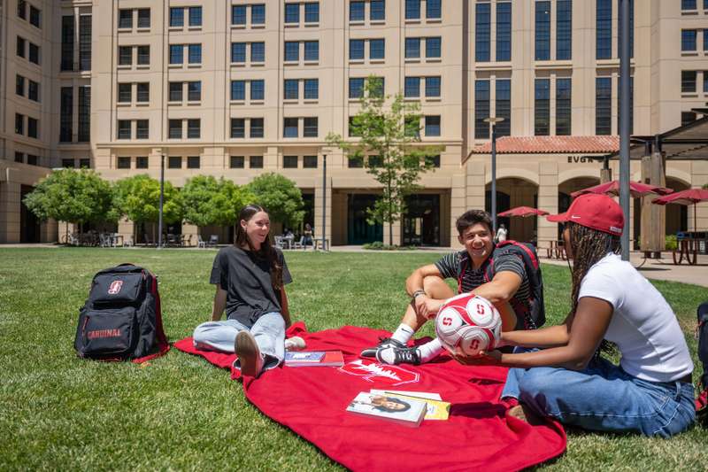 Students in front of a large dorm, sitting in the grass with a soccer ball