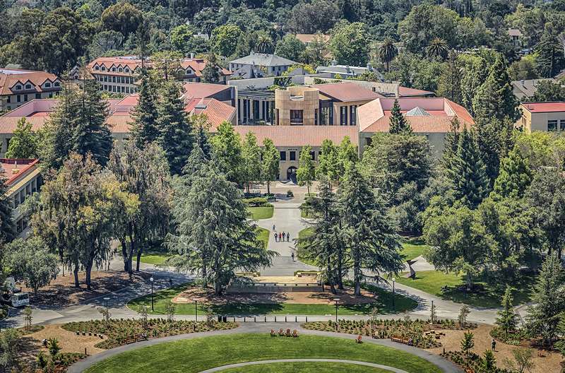 Aerial view of the Stanford campus, with the image centered on the Law School's William H. Neukom Building, with tree-lined pathways leading to historic red-roofed buildings.