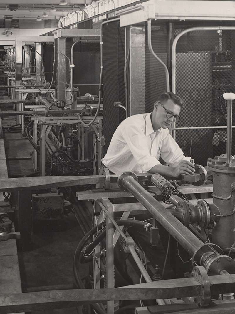 A man in a button-up shirt adjusts equipment along a long row of scientific machinery in a mid-20th century laboratory.