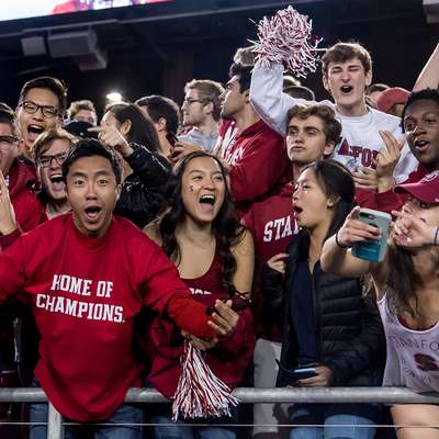 Enthusiastic Stanford fans in cardinal gear cheer from the stands at a football game.