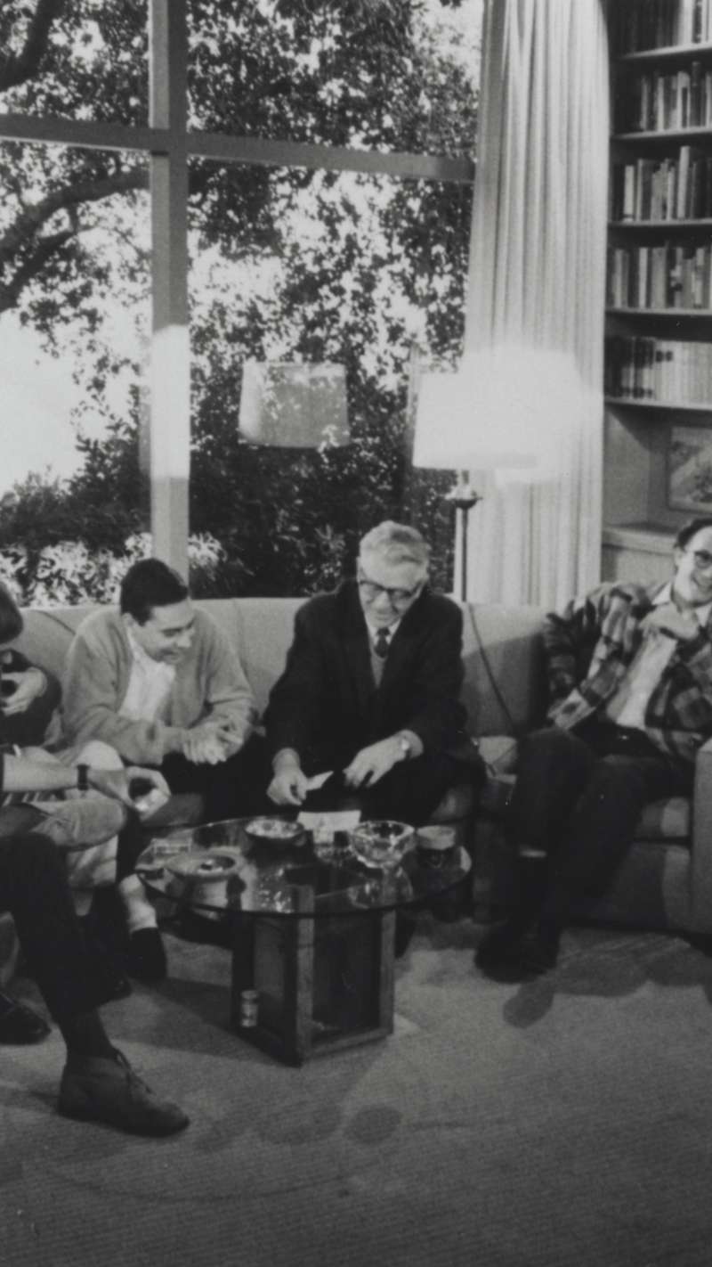 A group of people sits closely on a couch in a room on the Stanford campus, engaged in conversation, with a large window and bookshelf in the background.