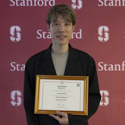 Kuniaki Iwanami holds a framed graduate certificate in artificial intelligence stands in front of a Stanford-themed backdrop.
