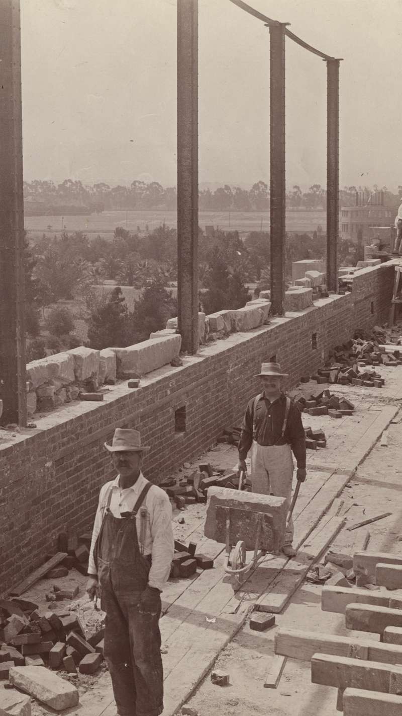 Two laborers work on the building site for Stanford men’s gymnasium in 1887, surrounded by construction materials and a distant view of trees and a building.