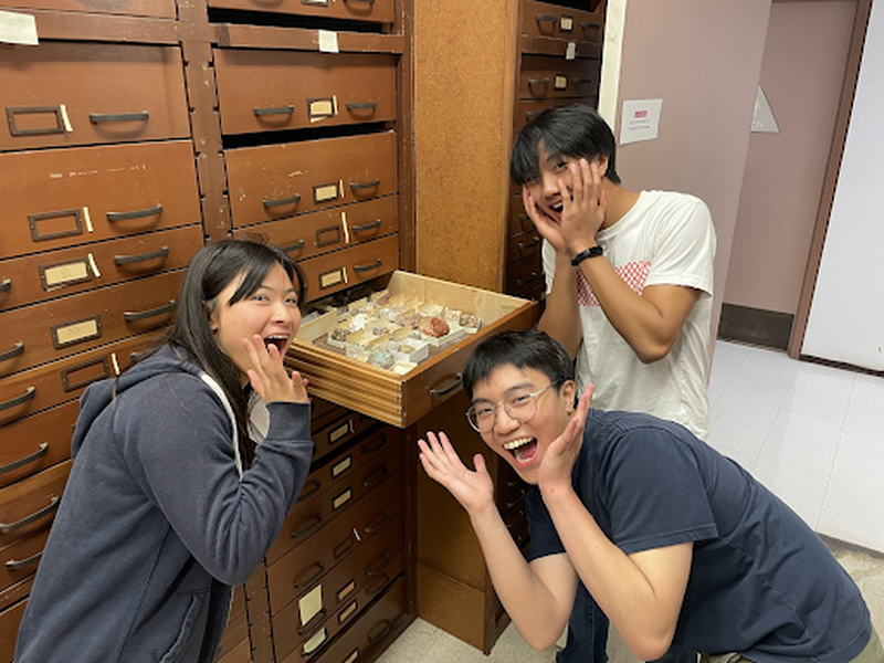 Three students pose playfully around a tray of mineral samples in a classroom or lab, showcasing their excitement.