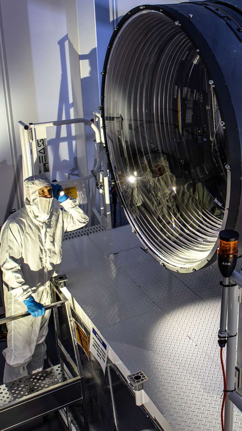 A technician in a white cleanroom suit stands beside a massive cylindrical telescope camera with a large lens, inspecting it with a flashlight in a lab.