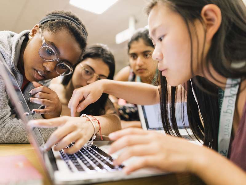 Students collaborate around a laptop, sharing ideas and pointing at the screen during a group project in a classroom setting.