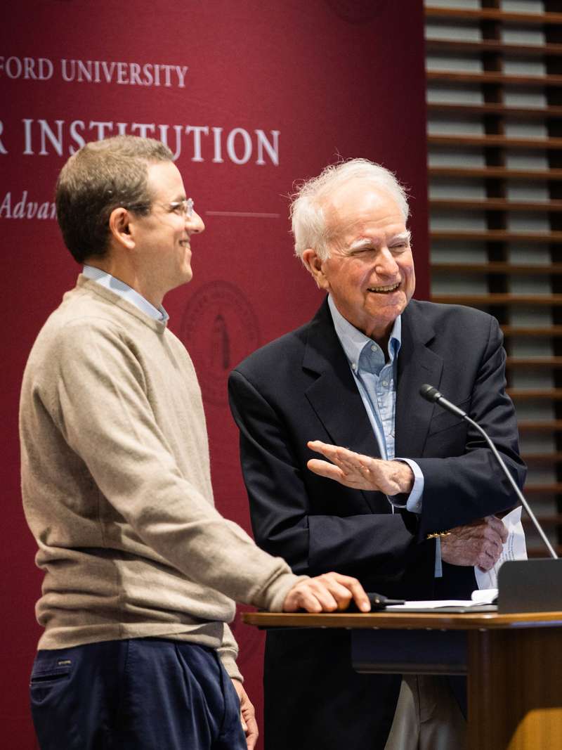 Robert E. Hall smiles at a podium in front of a backdrop that has the words "Stanford University" and "Hoover Institution" on it