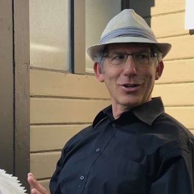 A man in a dark blue shirt and white hat sits near a door, gesturing with his hand as he engages in conversation.