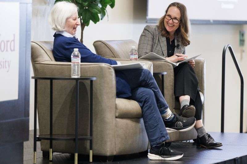 Two speakers sit in armchairs on stage, discussing topics while holding notes at a Stanford University event.