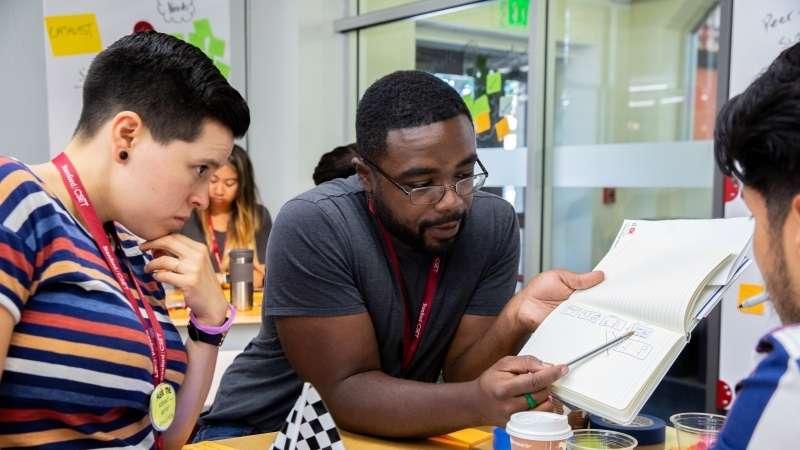 A group of adult students wearing red lanyards look at a prototyping sketch in a notebook