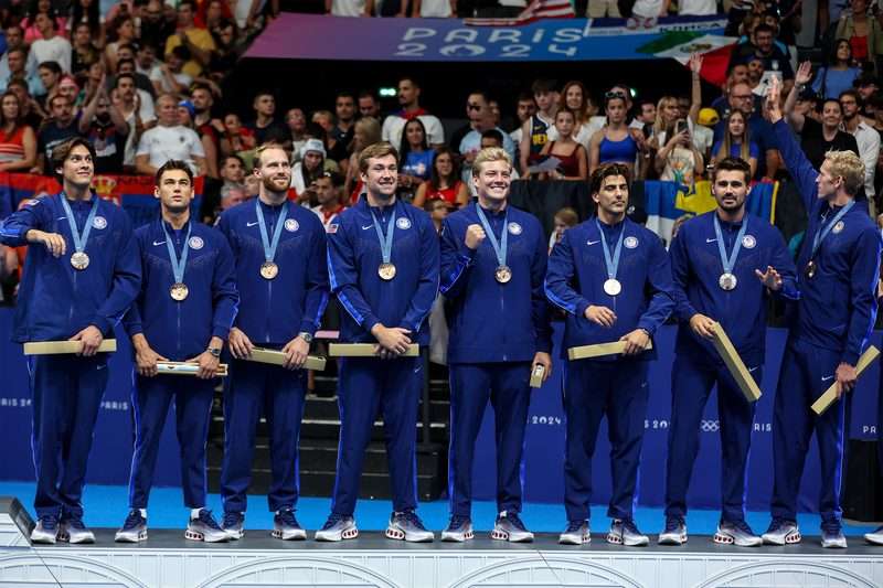 The US men's water polo team stands in a row in blue Team USA tracksuits wearing their bronze medals on a podium at the Paris 2024 Olympic Games.