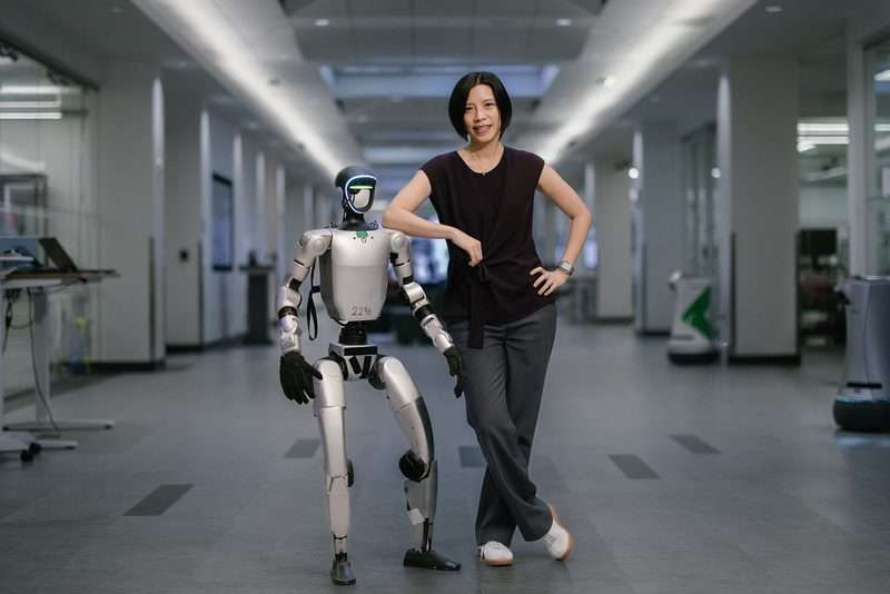 Karen Liu poses alongside a silver humanoid robot in a modern research lab, resting her arm on its shoulder.