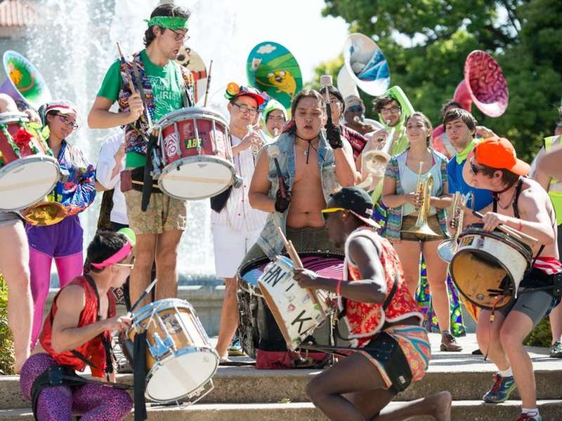 Members of a marching band in colorful, eclectic outfits energetically play drums, trumpets, and sousaphones near a fountain during an outdoor admit-weekend performance.