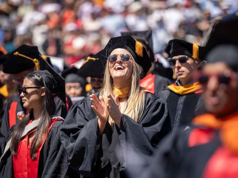 A smiling graduate in cap, gown, and sunglasses claps among a crowd of fellow graduates at an outdoor commencement ceremony on a sunny day.