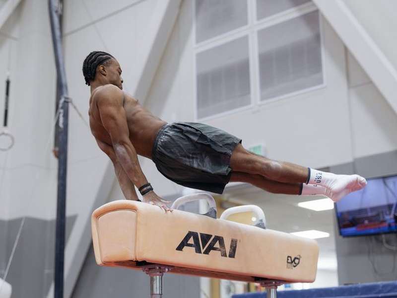 Gymnast Khoi Young performs a pommel horse routine, body extended horizontally and legs straight out, supporting himself on one arm in a training gym.