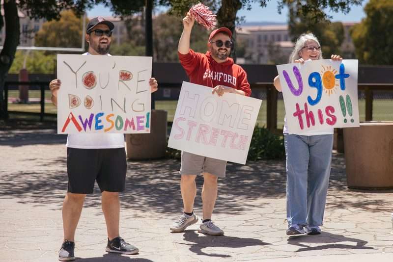 Three people hold handmade signs reading "You're doing awesome!", "Home stretch", and "U got this!!" cheering walkers along a path.
