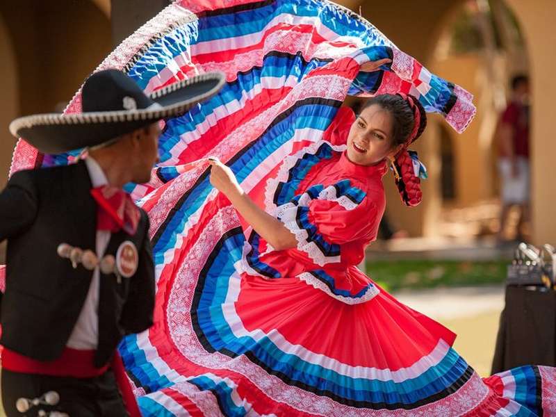 Two Ballet Folklórico dancers perform outdoors; a woman in a vivid red, white, and blue ruffled dress swirls her skirt beside a partner in a charro suit and sombrero.
