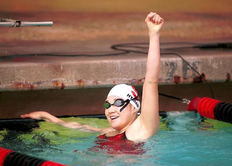 Misty Hyman raises her fist in celebration in the pool after a race, wearing a swim cap with her name and goggles, smiling.