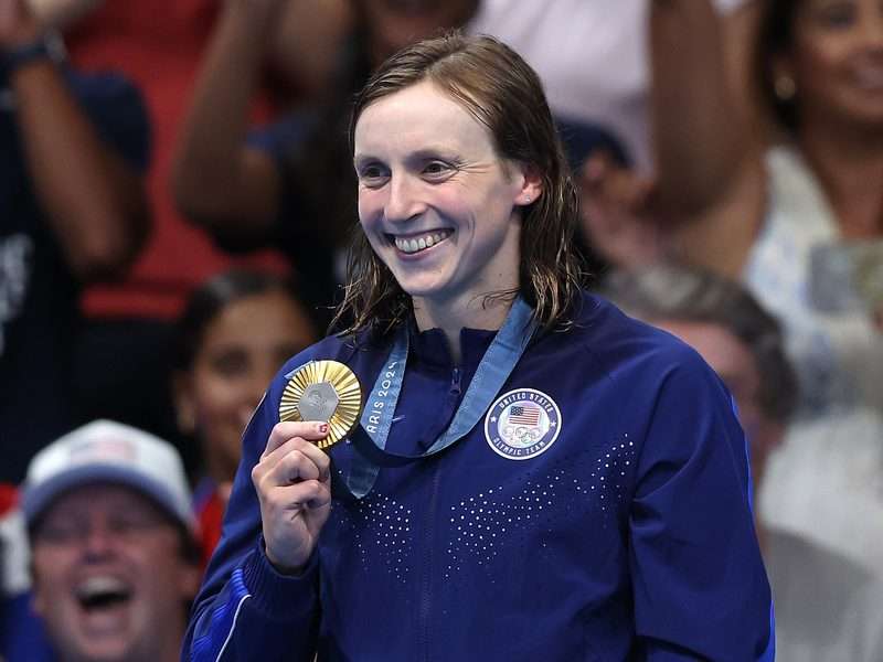 Katie Ledecky smiles and holds up her gold medal, wearing a blue Team USA jacket, in front of a cheering crowd at the Paris 2024 Olympic Games.