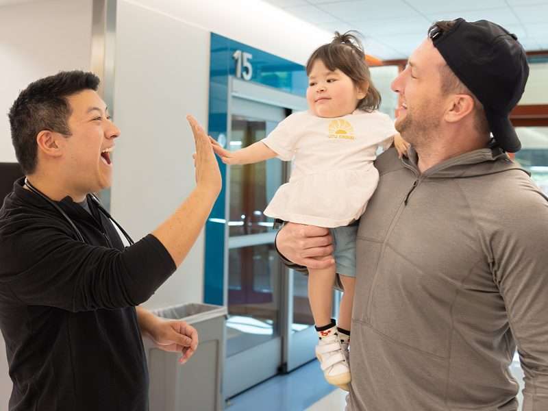 A dad holds his toddler girl, who is giving a high five to a man wearing a black shirt