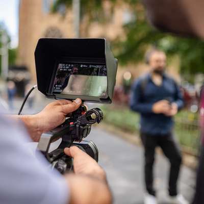 A camera operator films a man walking outdoors, seen through the video monitor in the foreground.