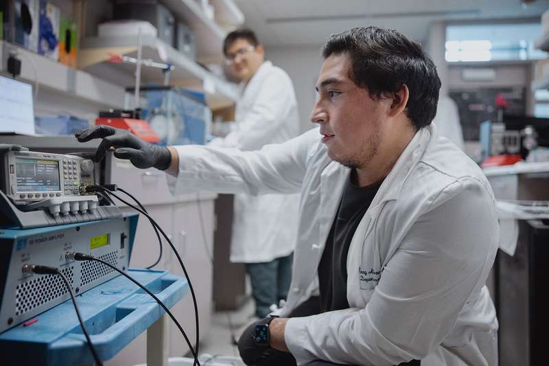 A researcher crouches to adjust equipment in a lab while a second researcher works at a bench in the background.