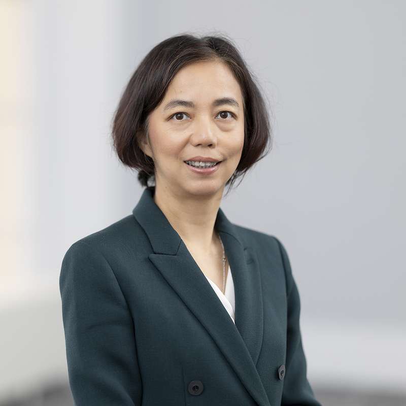 Fei-Fei Li wearing a dark green blazer over a white top, smiling against a blurred indoor background.
