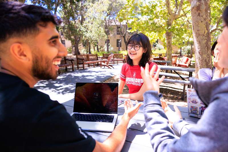 A group of students discussing and collaborating around a laptop at an outdoor table in a shaded area.