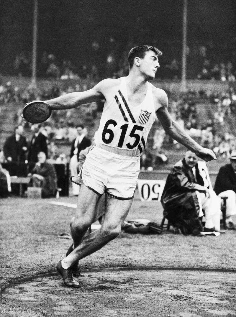 Bob Mathias, wearing a USA jersey numbered 615, winds up to throw the discus during a track and field competition, in this black-and-white historical photo.