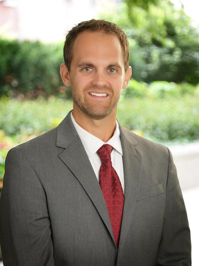 Smiling man in a gray suit with a red tie, standing outdoors with greenery in the background.
