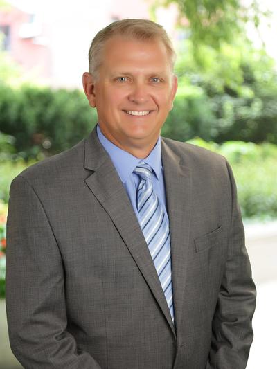 A smiling man in a gray suit and striped tie stands outdoors with lush greenery in the background.