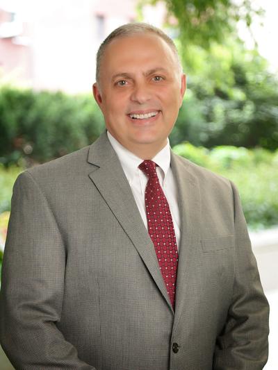 Charles Mehanna in a gray suit and red tie with greenery in the background.