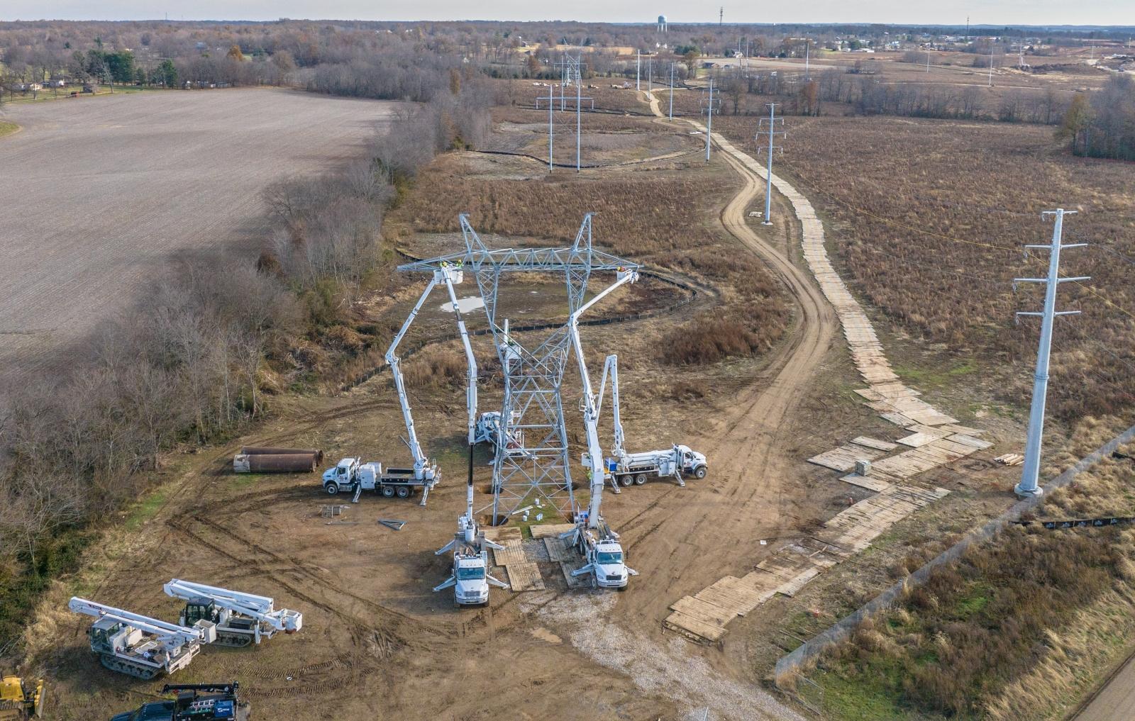 Construction site with multiple cranes and trucks assembling a large metal transmission tower in a rural landscape with dirt paths and fields