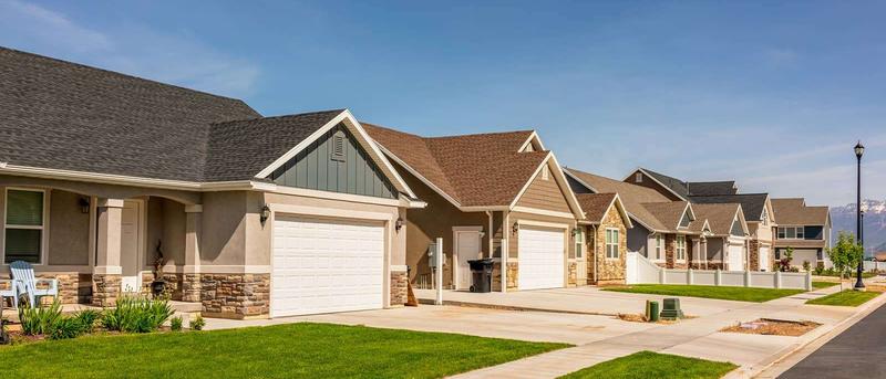 Row of suburban houses with neatly trimmed lawns, garages, and driveways under a clear blue sky.