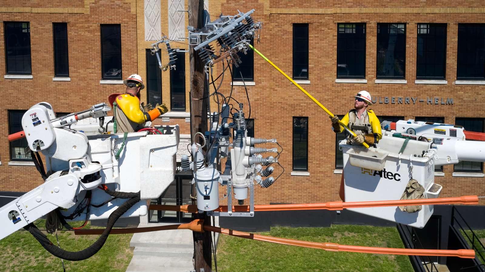 Two utility workers in bucket trucks repair power lines on a pole in front of a brick building. Both wear safety gear and helmets.