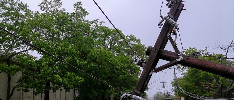 A leaning utility pole with tangled wires on a street, surrounded by greenery, under an overcast sky.