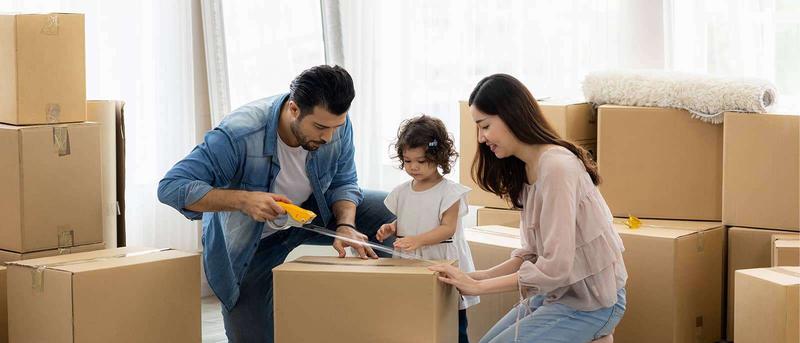 Family packing moving boxes, with a father sealing a box, a young child assisting, and a mother arranging. Brown boxes are stacked around them.