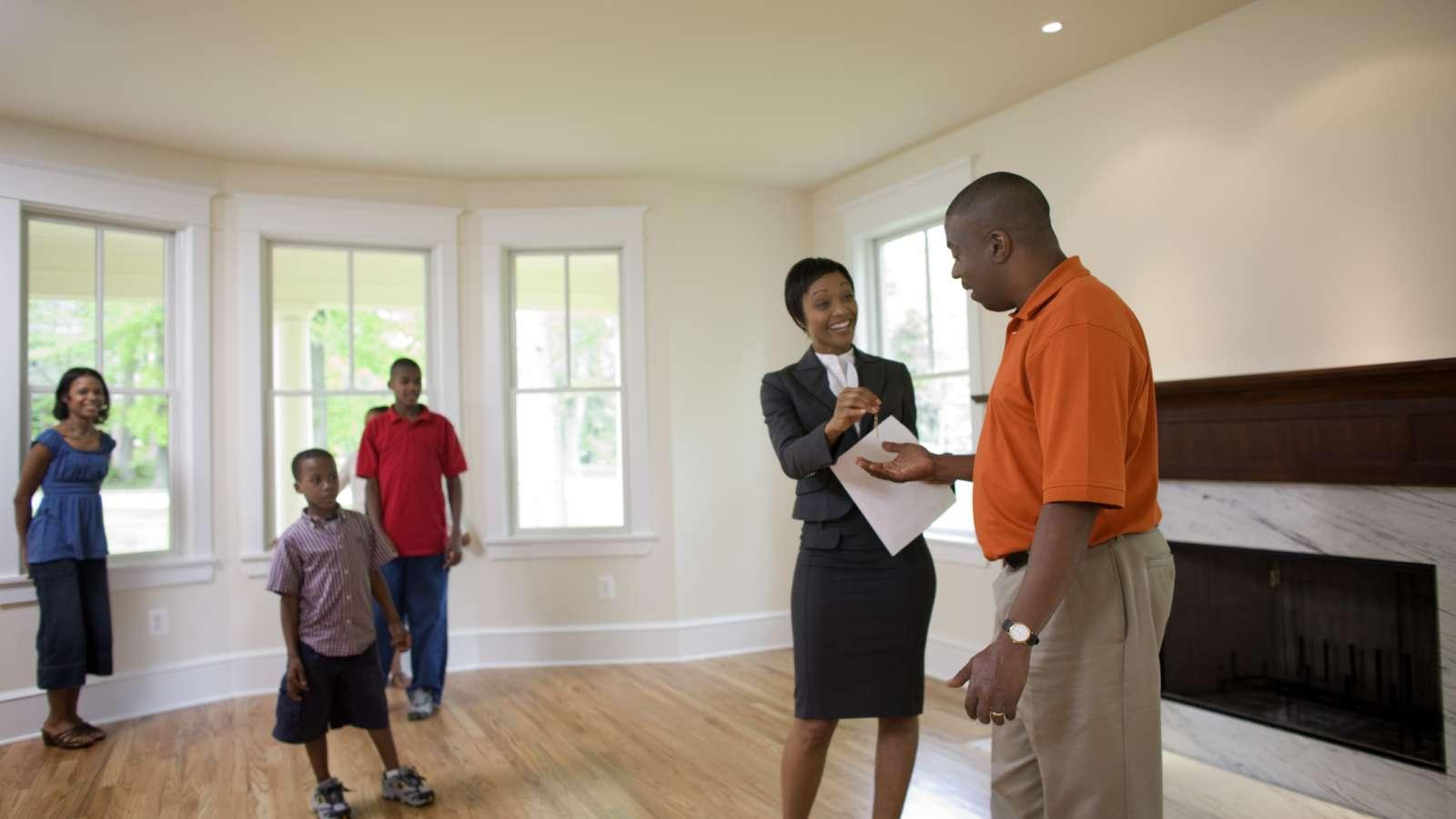 A real estate agent shows a family a bright living room with hardwood floors, large windows, and a fireplace.