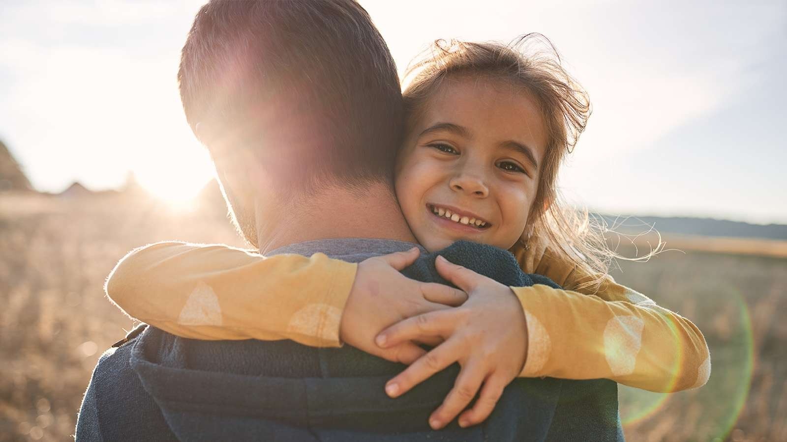 A young girl smiling and hugging a person in an outdoor setting, with sunlight creating a warm glow in the background.