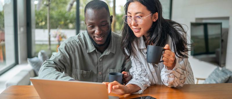 A man and woman smile while looking at a laptop. The woman holds a mug, and a tablet lies on the table in a bright, modern room.