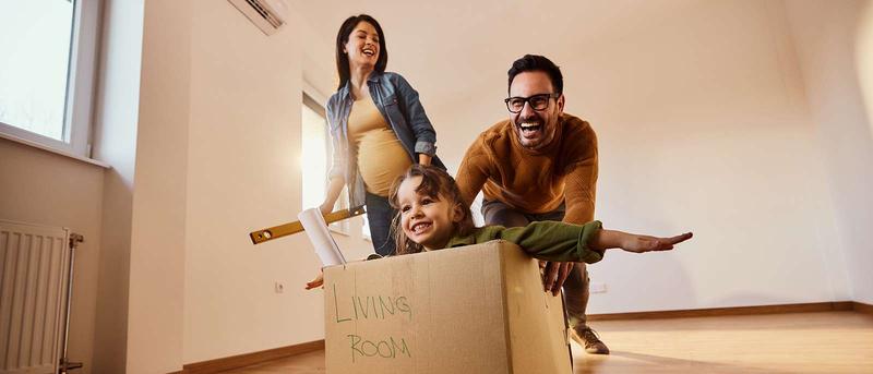 Family having fun in a new home, child sitting in a cardboard box labeled "Living Room #4," pushed by smiling parents in an empty room.