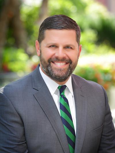 A man in a gray suit with a blue and green striped tie smiles, standing outside with greenery in the blurred background.