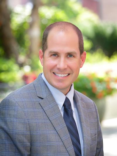 A man in a gray plaid suit and striped tie smiles outdoors, with greenery and colorful flowers in the blurred background.