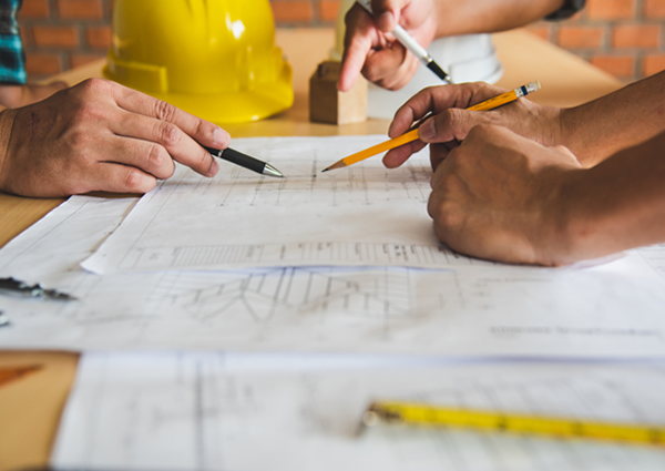 Hands drawing on building plan laid out on a desk.