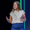 Smiling woman on stage at CommunityLIVE wearing a cream blouse and denim skirt, gesturing with hands and speaking into a headset microphone against a dark backdrop.
